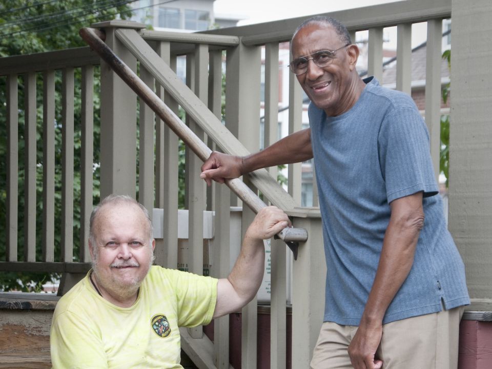 An older adult sits on steps as another stands at the bottom of the steps with his hand on the railing 